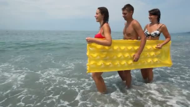 Dos mujeres y hombre sostiene el colchón inflable que camina en la playa — Vídeo de stock