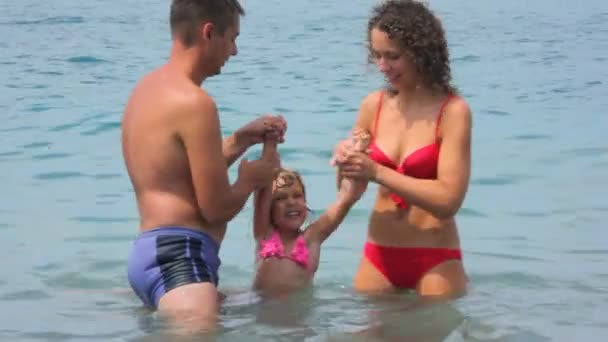 Homme et femme tient petite fille éclaboussant l'eau par les jambes dans la mer — Video
