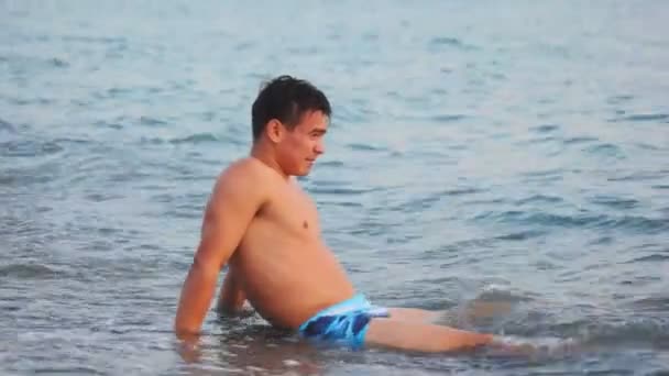 Young man sitting on rocky beach in front of surfing waves — Stock Video