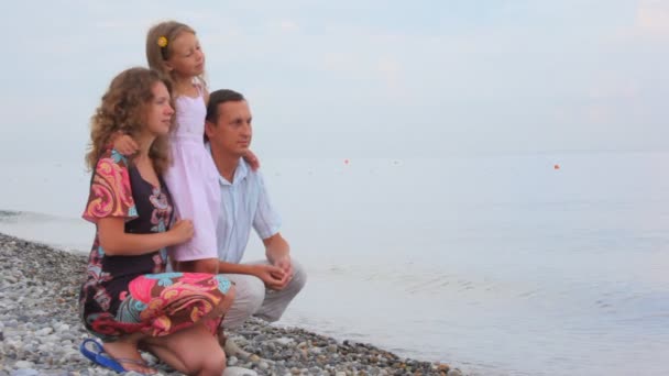 Family of three persons sitting on pebble coast and looks at sea — Stock Video