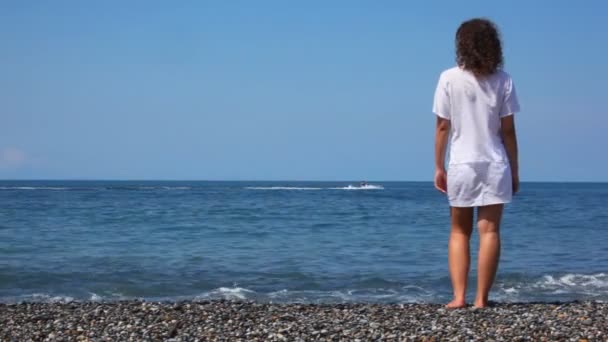 De pie mujer joven en la playa rocosa mira cortador en el mar — Vídeos de Stock