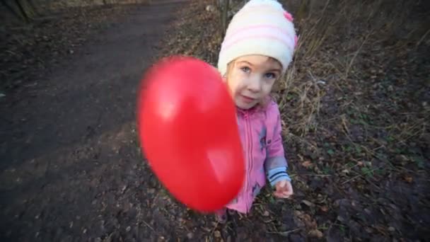 Niña sacudiendo globo rojo del corazón de pie en el bosque — Vídeo de stock