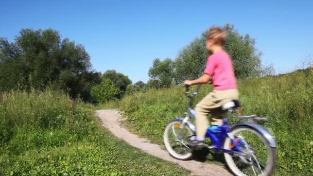 Маленький мальчик в розовом жилете едет вперед на голубом велосипеде в парке летом — стоковое видео