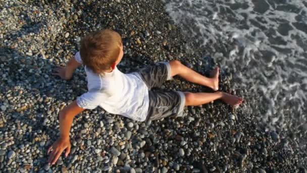 Young boy sits on pebble beach and looks at sea, top view — Stock Video