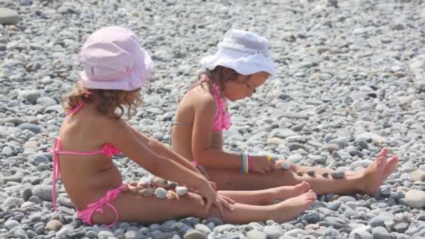 Two little girls sitting on stones and putting stones on their legs, side view — Stock Video