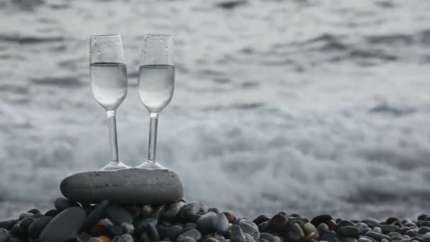 Two glasses with wine standing on stone on pebble beach, sea surf in background — Stock Video