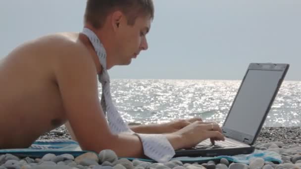 水泳トランクスとネクタイの海に対してノートブックとビーチに横たわってで服を着た男 — ストック動画