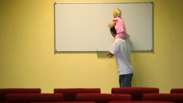 Hombre sosteniendo niña en hombros limpia pizarra blanca en sala de conferencias — Vídeo de stock