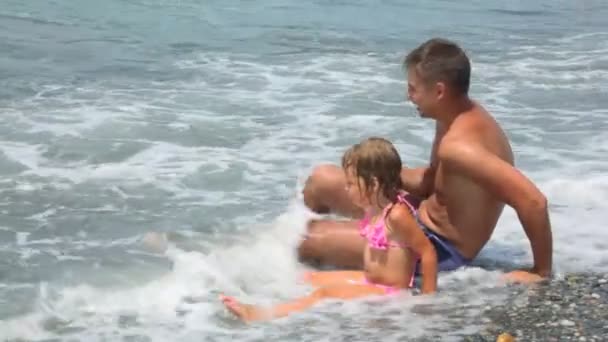 Uomo con la bambina si diverte si siede sulla spiaggia di ghiaia sotto le onde del mare surf — Video Stock