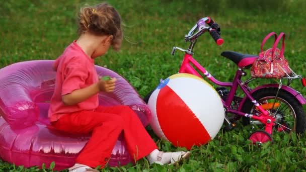 Chica sentarse en sillón inflable de los niños cerca de la bicicleta y la bola también mastica hojas — Vídeo de stock