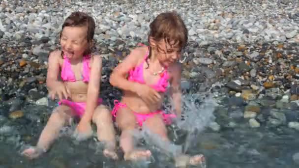 Två små flickor liggande på stenstrand med havet surfa och leka med vatten — Stockvideo