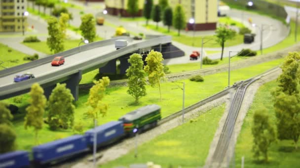Tren de juguete traer vagón de carga en el ferrocarril en la ciudad moderna de juguete entre la carretera, casa y árboles — Vídeo de stock
