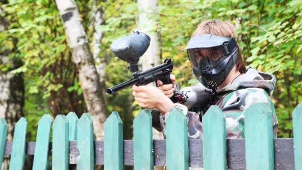 Homem de capacete e máscara com pistola de paintball se levanta para cerca e finge que dispara — Vídeo de Stock