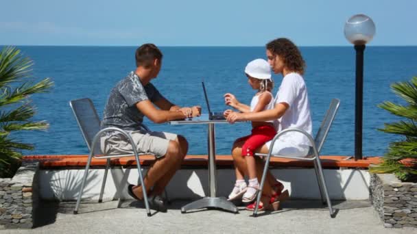 Family of three persons with notebook sits at table, sea in background — Stock Video