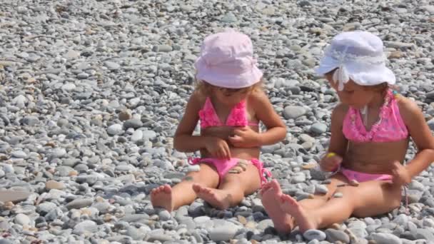 Two little girls sits on stones and puts stones on their legs, front view — Stock Video