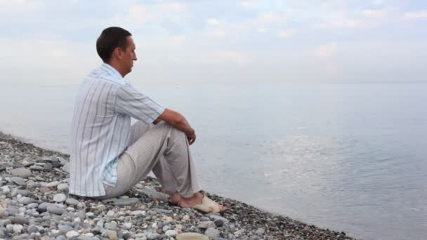 Hombre sentado en la costa de guijarros y mira al mar, vista lateral hacia atrás — Vídeos de Stock