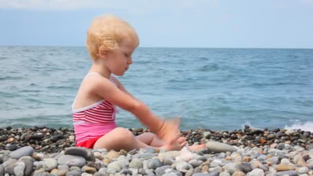 Niña sentada en la playa de guijarros y jugando con piedras, mar en el fondo — Vídeo de stock