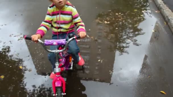 Маленькая девочка сидит на велосипеде на мокром асфальте с лужами, камера движется — стоковое видео