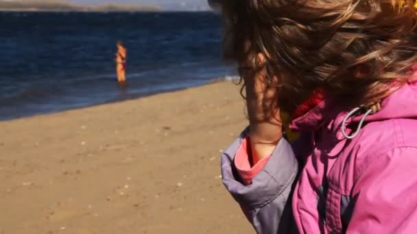 Menina senta-se na praia e olha para a mulher que se banha no rio — Vídeo de Stock