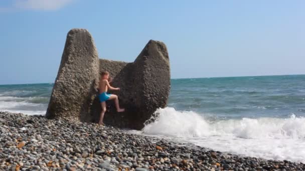 Мальчик прячется за валуном на пляже от морского серфинга — стоковое видео