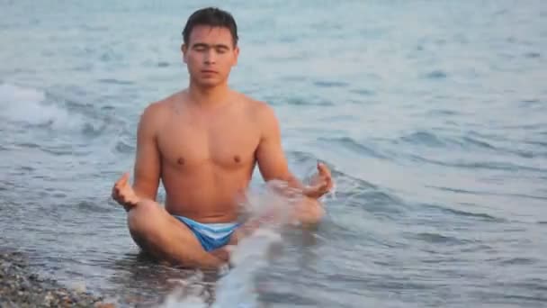 Mladý muž sedí v póze Lotus na skalnaté pláži s surfování vlny — Stock video
