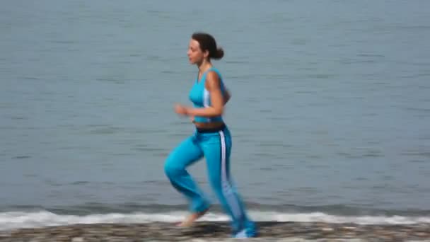 Mujer joven en chándal corriendo en la costa de guijarros, mar en el fondo — Vídeo de stock