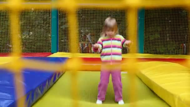 Little happy girl jumping on inflatable trampoline in amusement park behind net — Stock Video