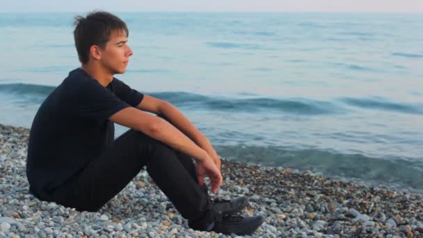 Teenager sits on pebble beach and looks at sea, profile — Stock Video