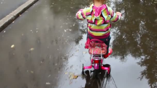 Niña pedaleando sobre asfalto mojado con charcos, la cámara la sigue — Vídeo de stock