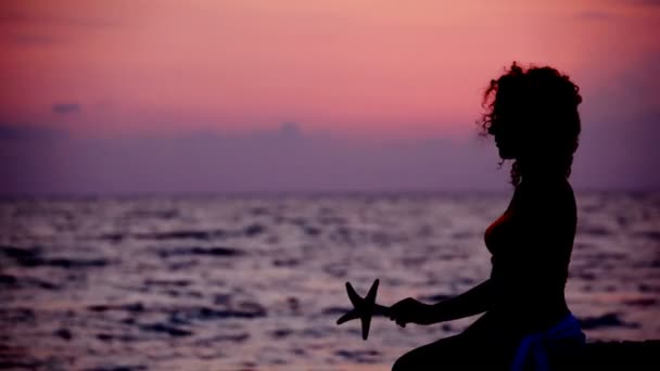 Silhouette di donna con stella di mare in mano sulla spiaggia, cielo di tramonto sullo sfondo — Video Stock