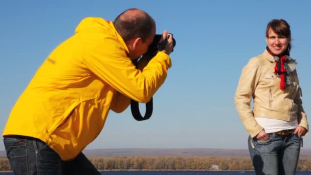 Мужчина фотографирует женщину на природе у реки, дерева и ясного неба — стоковое видео