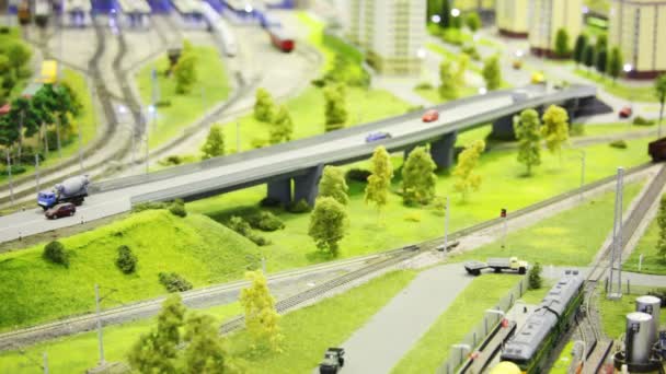 Tren de juguete empuja vagón de carga vacío en el ferrocarril en la ciudad moderna de juguete entre las carreteras, casa y árboles — Vídeos de Stock
