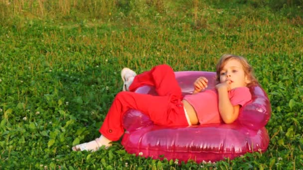 Flickan sitter i barnens uppblåsbara fåtölj, innehar finger i munnen och leende — Stock video