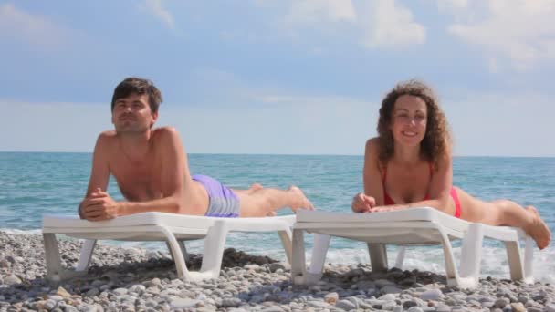 Happy couple lying on beach beds in pebble, clouds and sea in background — Stock Video