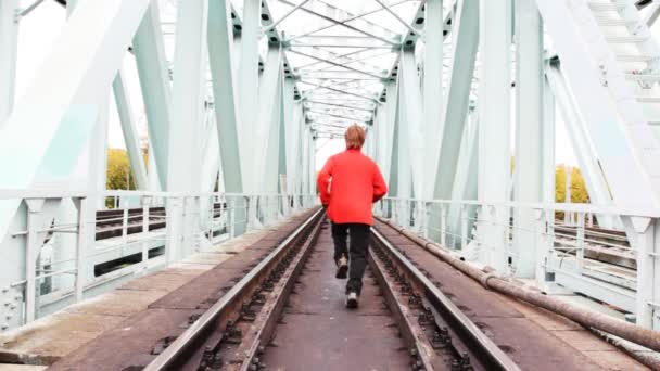 Rothaariger junger Mann läuft auf Eisenbahnbrücke vor Kamera zurück, hält dann an und wendet — Stockvideo