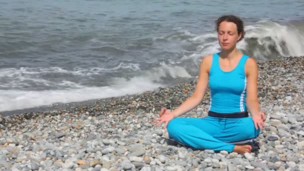 Jonge vrouw in trainingspak zit in pose van lotus op kiezelstrand tegen zee — Stockvideo