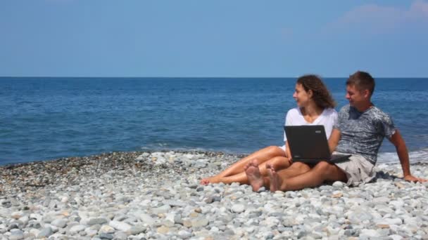 Happy woman and man with notebook sitting in rocky beach — Stock Video