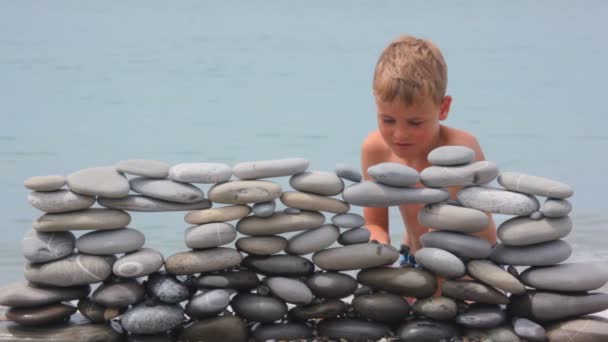 Niño jugando con la pared de piedras en la playa, mar surf en el fondo — Vídeos de Stock