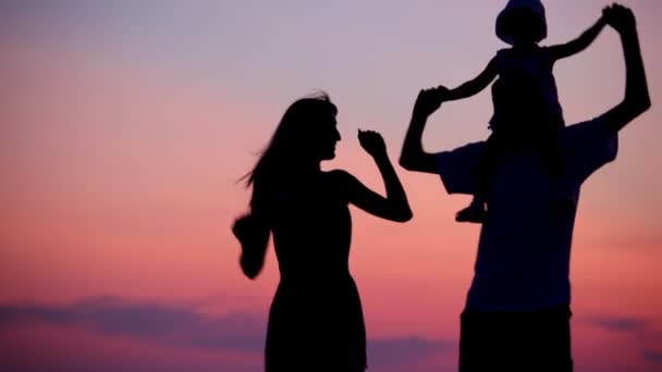 Женщина и мужчина держат маленькую девочку на плечах, танцуя на фоне заката — стоковое видео