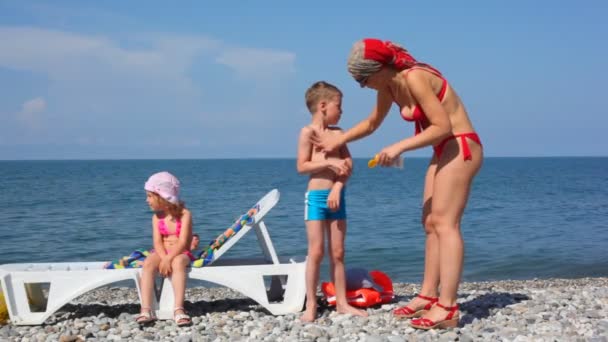 Familia en la playa de guijarros, madre extendiendo bronceador en hijo — Vídeos de Stock