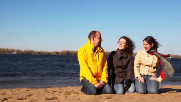 Man en twee vrouw zit op zand van de rivier en praten met elkaar — Stockvideo