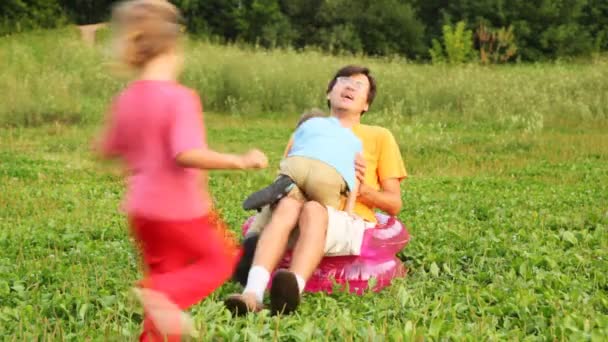 Mann, Junge und Mädchen sitzen in aufblasbarem Kindersessel auf Feld im Park und spielen den Narren — Stockvideo