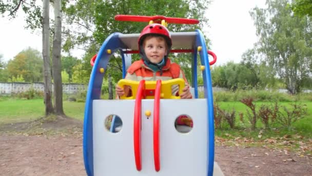 Menino em balanços de capacete em helicóptero de brinquedo no parque infantil — Vídeo de Stock