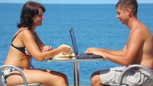 Женщина с сотовым телефоном и мужчина с помощью ноутбука сидит за столом, море в фоновом режиме — стоковое видео