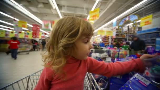 Niña sentada en el carrito de la compra de juguetes en el centro comercial — Vídeo de stock