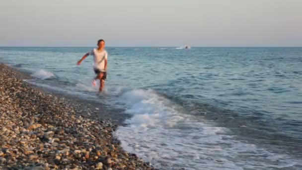 Мальчик бегает по галечному пляжу на камеру, морской серфинг на заднем плане — стоковое видео