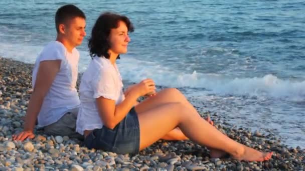 Junge Frau und Mann sitzen in Kiesstrand am Meer — Stockvideo