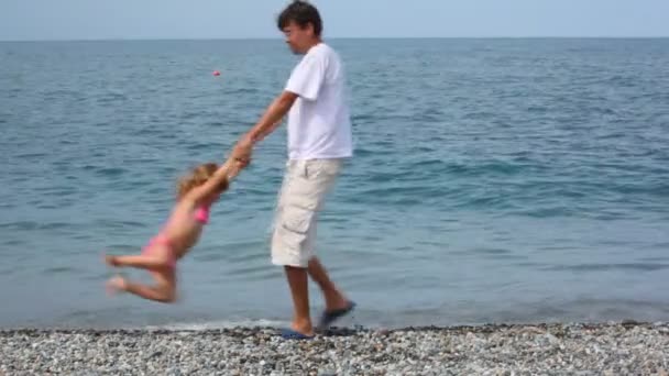 Man rotating little girl on pebble beach, sea in background — Stock Video