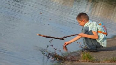 sırt çantası ile çocuk kıyısında oturur ve su yüzeyi üzerinde sopa ile dövmek