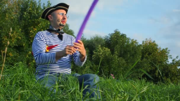 Man in piraat kostuum zit op gras in park en maakt zwaard uit luchtballon — Stockvideo